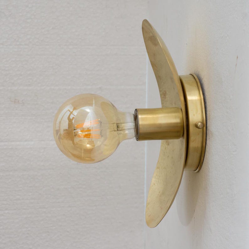 Puede incluir: Aplique de pared de lat&oacute;n con un dise&ntilde;o curvo decorativo. El aplique tiene una base redonda y un brazo cil&iacute;ndrico que sostiene una bombilla de estilo vintage. La bombilla emite un brillo &aacute;mbar c&aacute;lido, y el accesorio est&aacute; montado en una pared blanca.