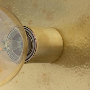 Puede incluir: Primer plano de una l&aacute;mpara de lat&oacute;n con una bombilla de cristal transparente. La l&aacute;mpara tiene una base cil&iacute;ndrica y est&aacute; montada sobre una superficie de lat&oacute;n texturizado. La bombilla est&aacute; encendida, emitiendo un brillo c&aacute;lido.