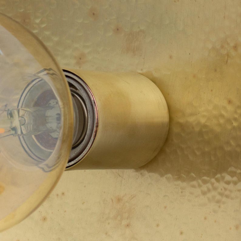 Puede incluir: Primer plano de una l&aacute;mpara de lat&oacute;n con una bombilla de cristal transparente. La l&aacute;mpara tiene una base cil&iacute;ndrica y est&aacute; montada sobre una superficie de lat&oacute;n texturizado. La bombilla est&aacute; encendida, emitiendo un brillo c&aacute;lido.