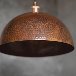 以下が含まれることがあります： ハンマー加工が施された銅色のペンダントライト。ドーム型のシェードは、銅色のステムとキャノピーから吊り下げられています。シェードの内側は、より濃い銅色です。背景はぼやけたグレーです。