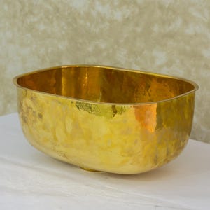 May include: A large, oval-shaped, hammered brass bowl with a smooth, shiny finish. The bowl is empty and sits on a white surface.