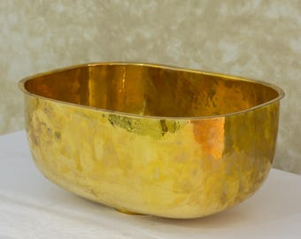 May include: A large, oval-shaped, hammered brass bowl with a smooth, shiny finish. The bowl is empty and sits on a white surface.