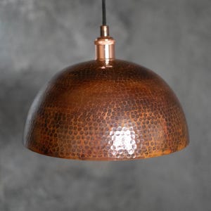 May include: A copper pendant light fixture with a hammered texture. The dome-shaped shade is suspended from a black cord and a copper-colored fixture. The light has a warm, metallic finish.