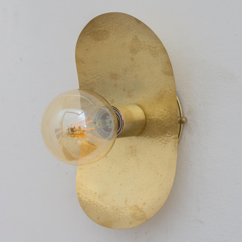 Puede incluir: Aplique de pared de lat&oacute;n con una placa trasera texturizada y asim&eacute;trica y una bombilla redonda expuesta. La bombilla es de color &aacute;mbar c&aacute;lido y la l&aacute;mpara est&aacute; montada en una pared blanca. El dise&ntilde;o es minimalista y moderno.