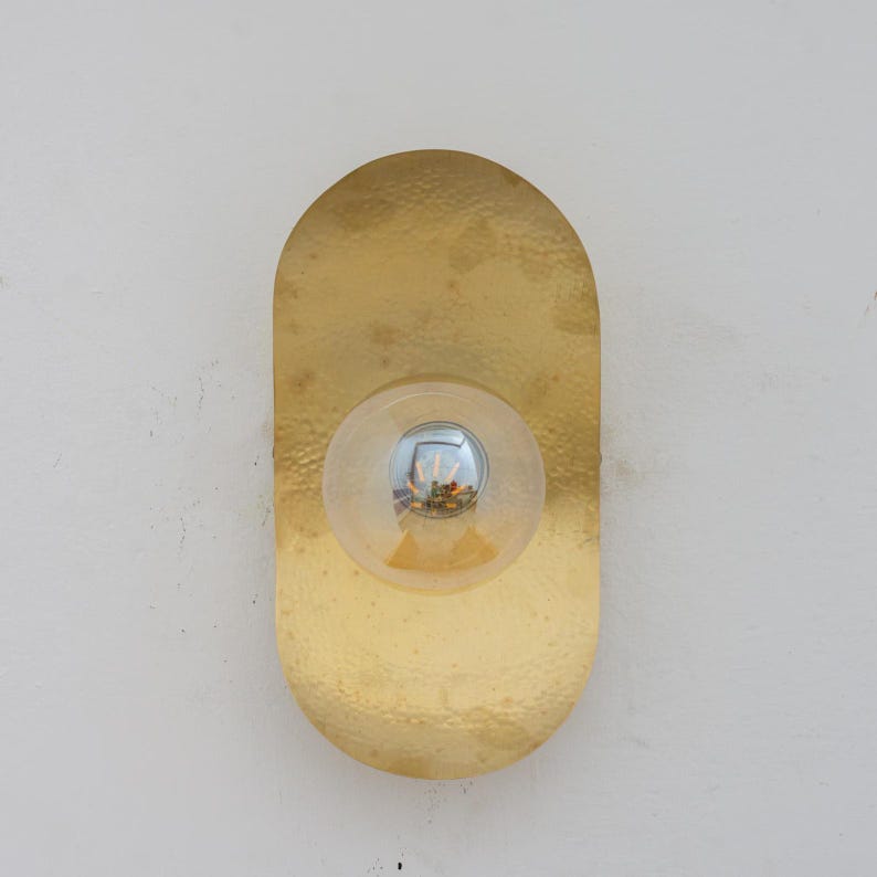 Puede incluir: Aplique de pared dorado con textura martillada y forma ovalada. El aplique presenta una bombilla redonda y transparente en el centro. La bombilla refleja el entorno.