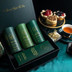 May include: A green gift box with a gold inscription "A Special Gift For You" contains four green tea canisters with gold lettering. The canisters are labeled "Earl Grey", "Yerba Mate Tea", "Pretty Peppermints", and "Tea Infuser Glass Bottle with Cup". The box is open and the canisters are visible. The box is sitting on a green surface with a cup of tea, a plate of desserts, and a chocolate cake.