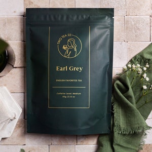 May include: A green pouch with gold lettering that reads "TWO TEA 22" and "Earl Grey" with the text "ENGLISH FAVORITES TEA" and "Caffeine Level: Medium 60g/2.12 oz".