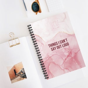 May include: A pink and white spiral bound notebook with a marbleized design and the text "Things I Can't Say Out Loud" in maroon.