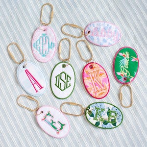 Pode incluir: Uma coleção de oito porta-chaves de tecido em formato oval, cada um com bordados, padrões e cores únicos. Os porta-chaves incluem vários monogramas, nomes e desenhos florais, todos presos a argolas de metal de tom dourado.
