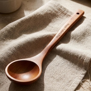 May include: A wooden ladle with a smooth, polished finish and a round bowl. The handle is engraved with the text "Happy Father's Day" and features a small hole at the end for hanging. The ladle rests on a beige, herringbone-patterned linen cloth.