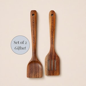 May include: A set of two wooden kitchen utensils, a solid spoon and a slotted turner, both featuring the engraved text COOK OFF CHAMP MMXXVI on the handle. A circular graphic to the left displays the text Set of 2 Giftset in black font.