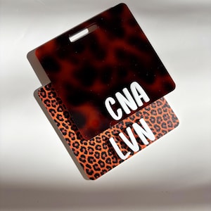 May include: Two rectangular ID badge holders. One has a tortoiseshell pattern with the letters "CNA" in white. The other has an orange and black leopard print with the letters "LVN" in white. Both have a slot for a lanyard.