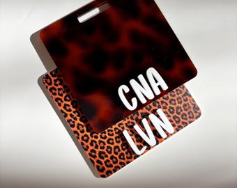 May include: Two rectangular ID badge holders. One has a tortoiseshell pattern with the letters "CNA" in white. The other has an orange and black leopard print with the letters "LVN" in white. Both have a slot for a lanyard.