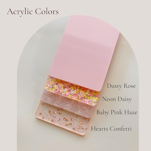 May include: A selection of acrylic colour samples in pink hues, labelled "Acrylic Colors." The samples include "Dusty Rose," "Neon Daisy," "Baby Pink Haze," and "Hearts Confetti," displaying different textures and glitter effects.