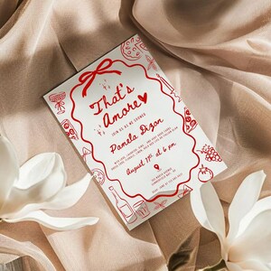 以下が含まれることがあります： 「That's Amore」というフレーズとパーティーの詳細が記載された、赤い文字とイラストが描かれた白い招待状。カードは食べ物に関連する落書きと赤いリボンで飾られています。カードは白い花が添えられたベージュの布の上に置かれています。