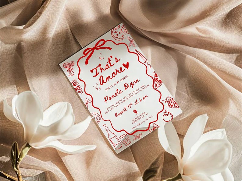 以下が含まれることがあります： 「That's Amore」というフレーズとパーティーの詳細が記載された、赤い文字とイラストが描かれた白い招待状。カードは食べ物に関連する落書きと赤いリボンで飾られています。カードは白い花が添えられたベージュの布の上に置かれています。