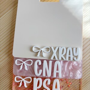 May include: A layered badge holder with the words "XRAY", "CNA", and "PSA" in white, with bow accents. The top layer is off-white, the middle layer is pink, and the bottom layer is clear with glitter.