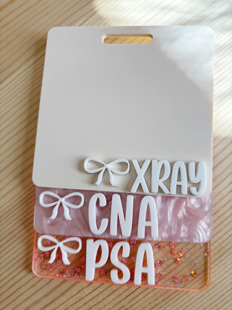 May include: A layered badge holder with the words "XRAY", "CNA", and "PSA" in white, with bow accents. The top layer is off-white, the middle layer is pink, and the bottom layer is clear with glitter.