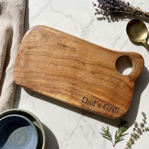May include: A rectangular wooden cutting board with a natural grain pattern and a circular handle hole. The board features the engraved text "Dad's Grill" in the bottom right corner. The wood has a warm brown tone with visible knots.