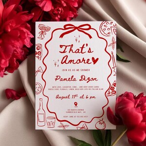 以下が含まれることがあります： 「That's Amore」というフレーズとイベントの詳細が記載された、赤い文字とイラストが特徴の白い招待状。カードは赤い花に囲まれ、クリーム色の布の上に置かれています。