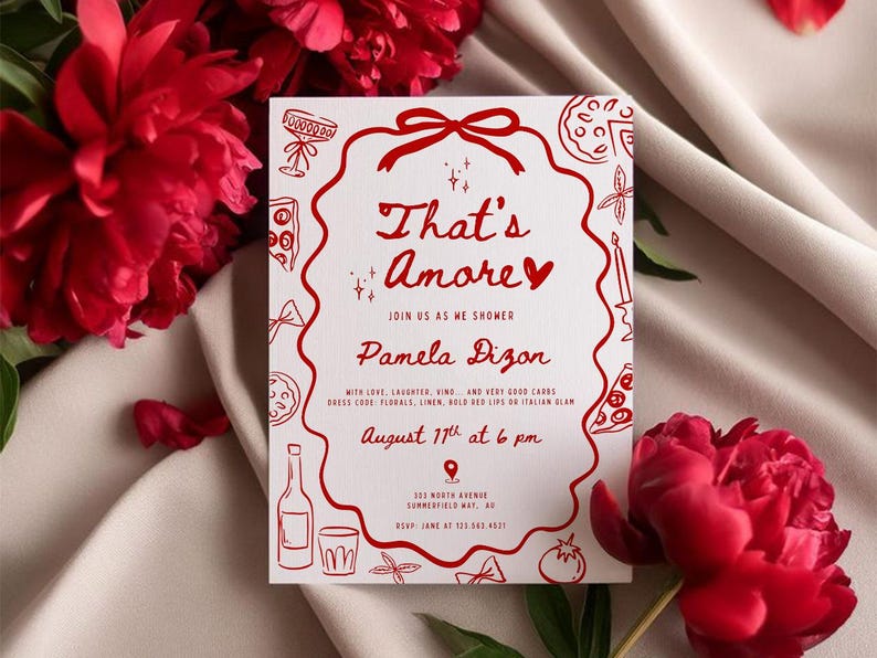 以下が含まれることがあります： 「That's Amore」というフレーズとイベントの詳細が記載された、赤い文字とイラストが特徴の白い招待状。カードは赤い花に囲まれ、クリーム色の布の上に置かれています。