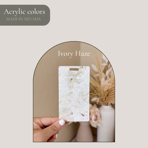 May include: A rectangular acrylic tag in the "Ivory Haze" colour, held by a hand. The tag has a hole at the top and a swirling, marbled pattern in shades of white and cream. The text "Acrylic colors MADE IN NEVADA" is in the upper left.