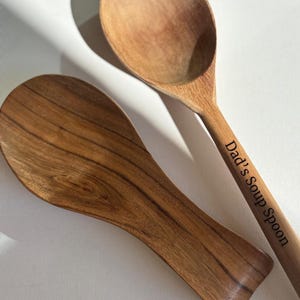 May include: A wooden spoon rest and a matching wooden spoon with the text Dad's Soup Spoon engraved on the handle. Both items feature a natural brown wood grain pattern with a smooth finish, arranged diagonally against a plain white background.