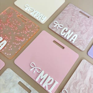May include: A selection of colourful, square name badges with a handle slot. The badges are in various colours, including pink, peach, and marbled patterns. Each badge has white text and a bow accent, with text such as "XRAY" and "MRI".