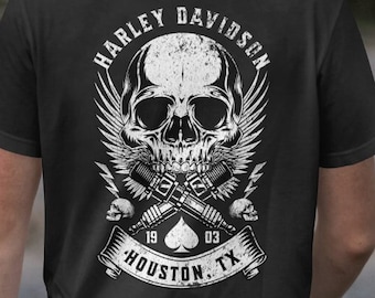 May include: Black t-shirt featuring a white graphic of a skull with wings, crossed tattoo guns, and the text "HARLEY DAVIDSON" above the skull. Below the skull is the text "HOUSTON, TX".