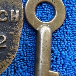 May include: Close-up of a vintage brass key with a circular top and a rectangular body. The top of the key has the letters "O.S.L." engraved. The key is set against a textured blue background, and a portion of a circular object with the letters "CH" and the number "2" is visible.