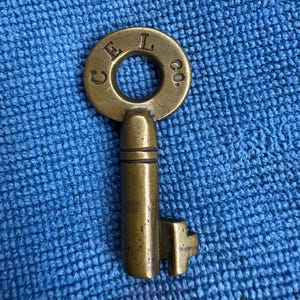 May include: A vintage brass key with a circular head and a rectangular bit. The head of the key has the text "CEL CO." engraved around the central hole. The key is set against a blue textured background.