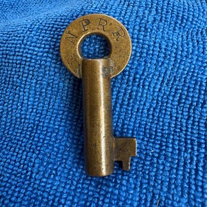 May include: A brass key with a round head that says "NPR" on it. The key has a long, straight shaft and a small, rectangular bit at the end.