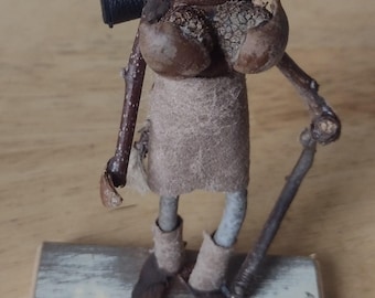 May include: A whimsical, handcrafted figurine of a woodland creature. It features a head and body made from natural materials, wearing a hat and carrying a walking stick. The figure stands on a small wooden base, showcasing rustic charm.