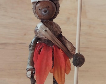 May include: A whimsical figurine made from natural materials, featuring a stick figure with a crown of orange and red fabric, holding a stick with a flower on top. The figurine is standing on a wooden base.