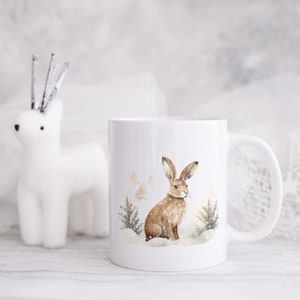 May include: White ceramic mug with a brown hare illustration in a snowy forest setting. The hare is sitting in the snow with trees behind it.