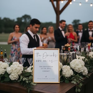 May include: A gold-framed sign with the text "The Drinks are on us" and "The Hangover is on you! Cheers!" sits on a wooden bar decorated with white flowers and greenery. A bartender is visible behind the bar, with guests in the background.