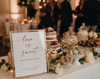 May include: A gold-framed sign on a white tablecloth reads love is sweet PLEASE TAKE A TREAT STEPHEN and KELLEY October 11, 2026. Beside it sits a tiered cake, glass jars of sweets, bowls of macarons, and white roses with green foliage.