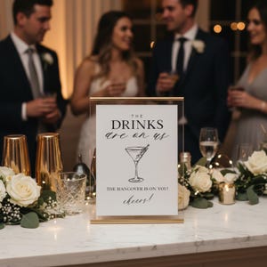 May include: A gold-framed sign with the text "THE DRINKS are on us" and "THE HANGOVER IS ON YOU! cheers!" A martini glass illustration is below the text. The sign is on a white marble table with floral arrangements and people in the background.