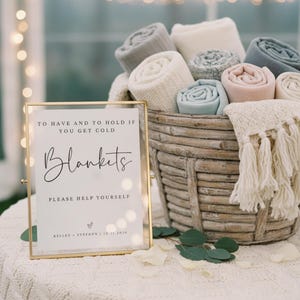 May include: A rustic woven basket filled with rolled blankets in various neutral colors, accompanied by a gold-framed sign that reads: TO HAVE AND TO HOLD IF YOU GET COLD, Blankets, PLEASE HELP YOURSELF, KELLEY + STEPHEN | 10.11.2026.