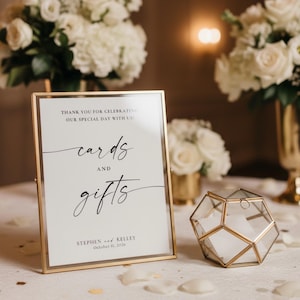May include: A vertical rectangular gold-framed sign with white background and black text reading "THANK YOU FOR CELEBRATING OUR SPECIAL DAY WITH US! cards AND gifts STEPHEN and KELLEY October 11, 2026" next to a geometric gold-framed glass box.