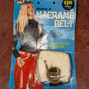 May include: Vintage macrame belt kit on a blue and white card. The kit includes macrame cord, leather thong, wooden beads, and working picks. The packaging features a woman wearing a macrame belt and the text "Make a Macrame Belt".