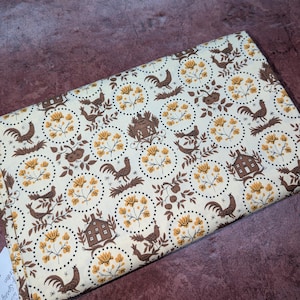K&ouml;nnte beinhalten: Cremefarbener Stoff mit einem sich wiederholenden Muster aus braunen H&auml;hnen, H&auml;usern und floralen Designs. Das Design ist in gepunkteten Kreisen angeordnet, mit gelben und braunen Akzenten. Der Stoff ist gefaltet und zeigt seine Textur und sein Muster.