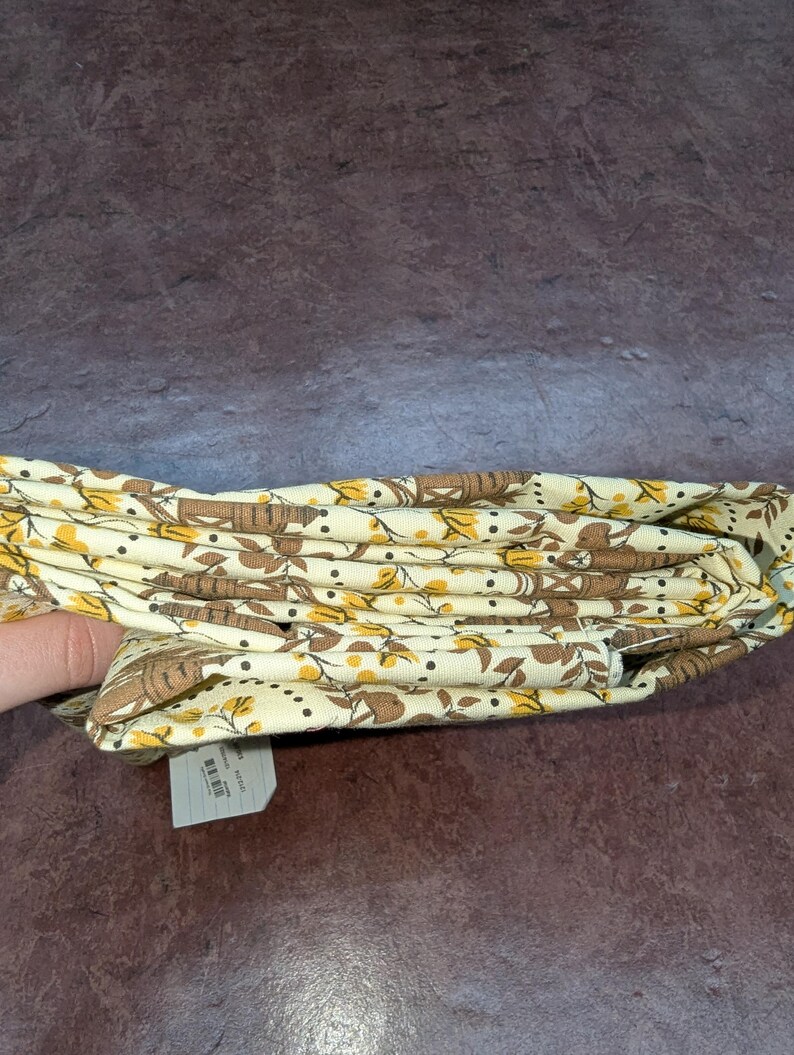 K&ouml;nnte beinhalten: Gefalteter Stoff mit cremefarbenem Hintergrund und einem sich wiederholenden Muster aus braunen baumartigen Formen und gelben Akzenten. Der Stoff ist gefaltet und zeigt das Muster. Ein wei&szlig;es Etikett ist sichtbar.