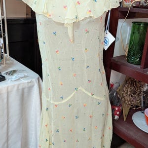May include: A vintage, pale yellow dress with a V-neck and ruffled neckline. The dress is adorned with small, colorful floral embroidery. The dress is displayed on a mannequin. A tag is attached to the dress.