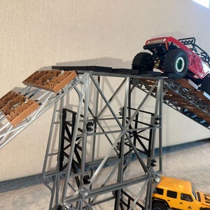 May include: A red and black toy truck driving up a ramp made of brown plastic pieces. The ramp is attached to a metal frame. A yellow toy truck is parked at the bottom of the ramp.