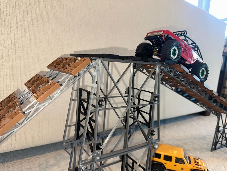 May include: A red and black toy truck driving up a ramp made of brown plastic pieces. The ramp is attached to a metal frame. A yellow toy truck is parked at the bottom of the ramp.