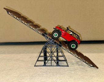 May include: A red toy off-road vehicle climbs a brown and grey ramp structure. The ramp is supported by a grey metal frame. The vehicle has black tyres with green accents.
