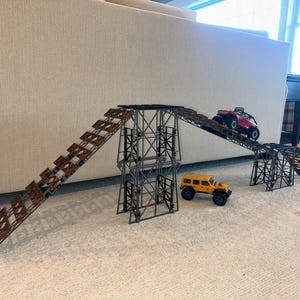 May include: A set of two metal ramps with wooden steps for toy cars. The ramps are connected by a metal bridge. A red toy car is driving up one ramp and a yellow toy car is driving down the other ramp.