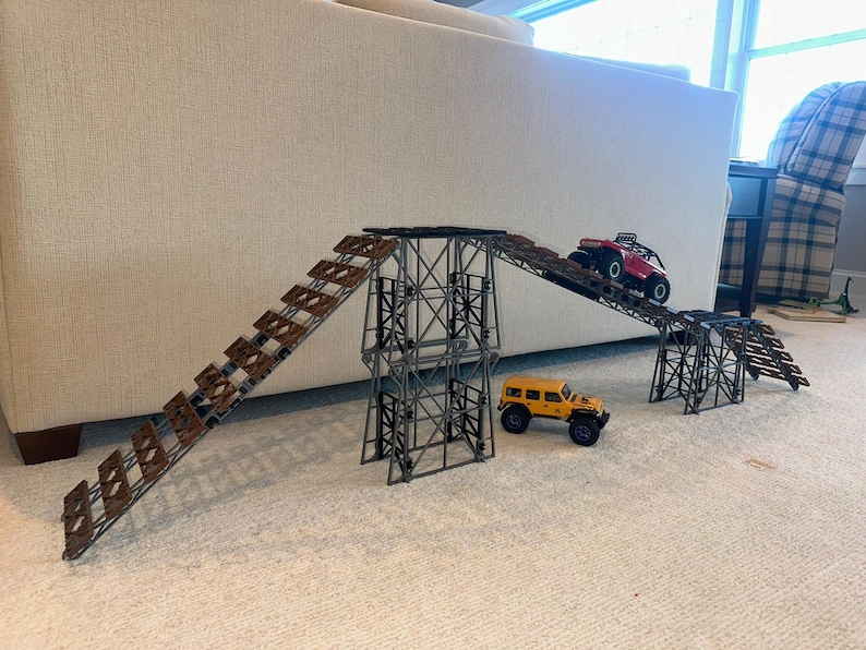 May include: A set of two metal ramps with wooden steps for toy cars. The ramps are connected by a metal bridge. A red toy car is driving up one ramp and a yellow toy car is driving down the other ramp.