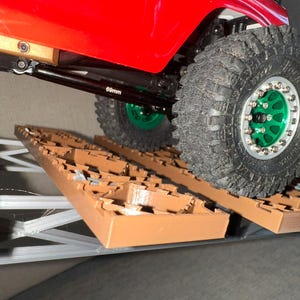 May include: A red toy truck with black axles and green wheels is driving over a brown Lego-like obstacle course. The truck has a 60mm axle.
